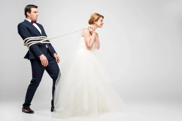 Bride in wedding dress pulling groom bound with rope, isolated on grey, feminism concept — Stock Photo