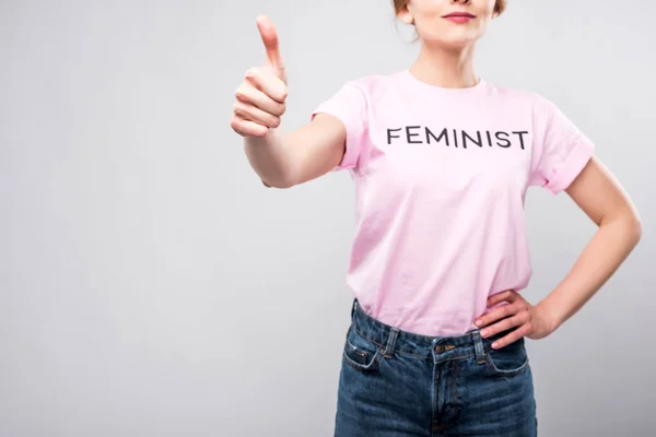 Vista recortada de la mujer en camiseta feminista rosa que muestra el pulgar hacia arriba, aislado en gris - foto de stock