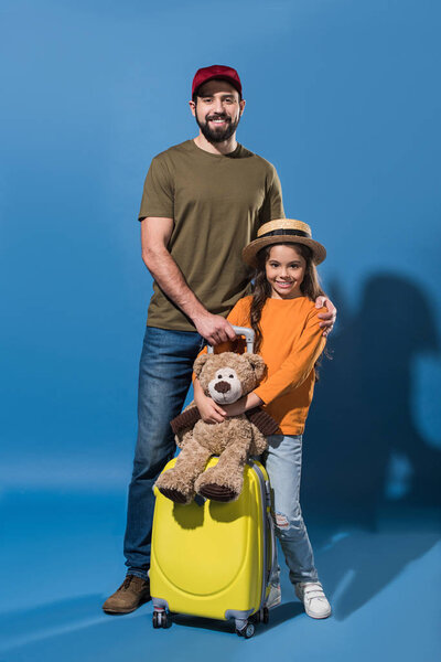 father in cap and daughter in straw hat standing with bag on wheels on blue