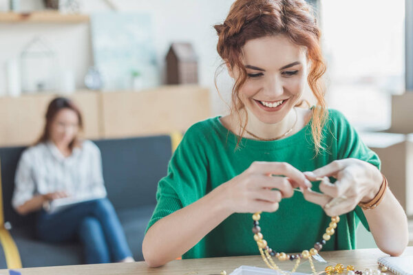 счастливая молодая женщина делает ожерелье из бусин в мастерской в то время как ее коллега сидит на диване размыто на заднем плане
