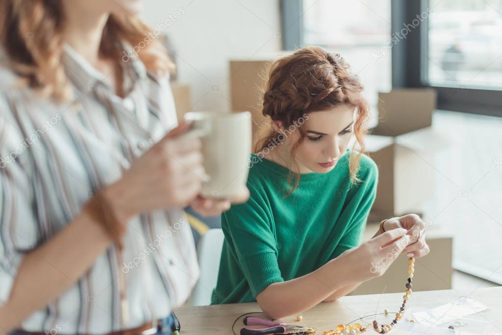 concentrated young woman making accessories in workshop while colleague drinking coffee on foreground