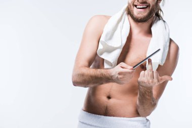 Smiling young man with bath towel around his neck holding nail file in hand and showing middle finger by another one,  isolated on white  clipart