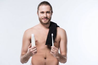 Cheerful young man with black towel on shoulder holding toothpaste in hand and toothbrush in another one,  isolated on white  clipart