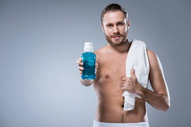 Banyo havlusu uzanmış el diş durulama ile omuz holding şişe üzerinde olan adam gülümsüyor, gri izole 