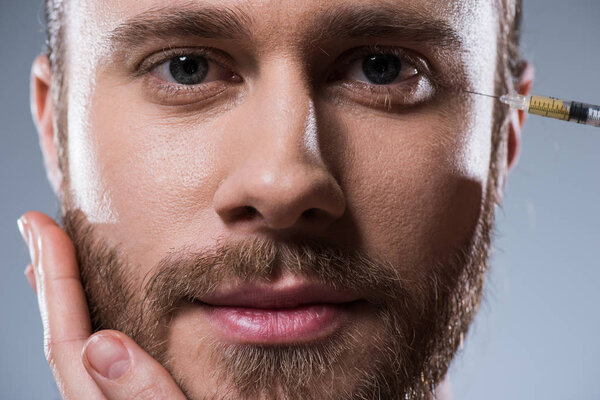 Портрет бородатого кавказца, смотрящего в камеру, в то время как лица, держащие его за подбородок и делающие укол, изолированы на сером
 