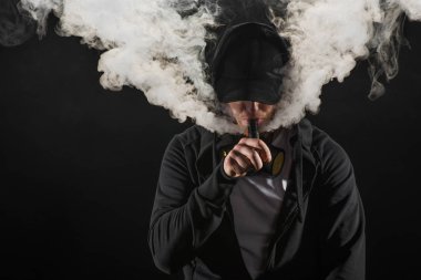 Young bearded man wearing cap exhaling smoke of electronic cigarette isolated on black clipart