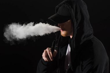 Young bearded man wearing cap exhaling smoke of electronic cigarette isolated on black clipart