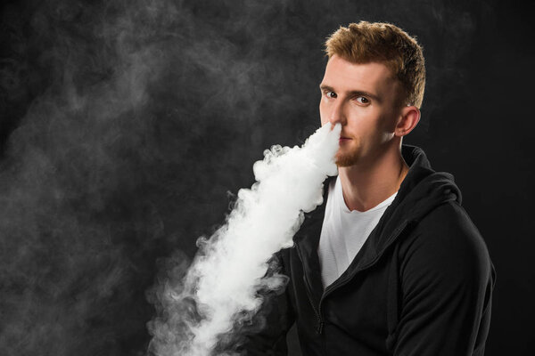 Young bearded man exhaling smoke of electronic cigarette surrounded by clouds of steam