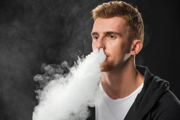 Young bearded man exhaling smoke of electronic cigarette surrounded by clouds of steam