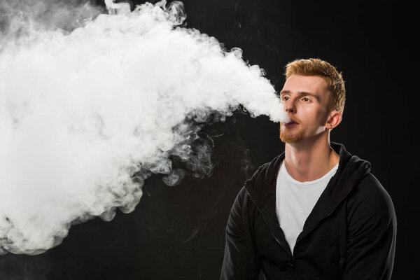 Young bearded man exhaling smoke of electronic cigarette surrounded by clouds of steam