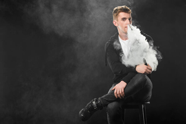 Young bearded man exhaling smoke of electronic cigarette surrounded by clouds of steam