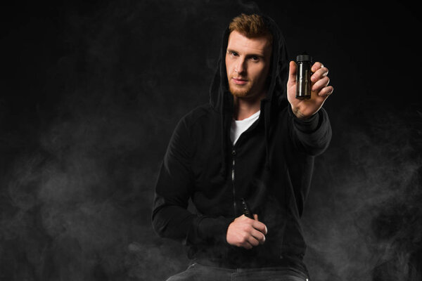 Young bearded man showing e-liquid bottle surrounded by clouds of steam