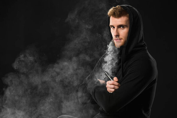 Young bearded man exhaling smoke of electronic cigarette surrounded by clouds of steam