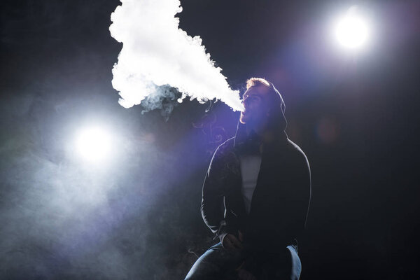 Young bearded man exhaling smoke of electronic cigarette against backlit