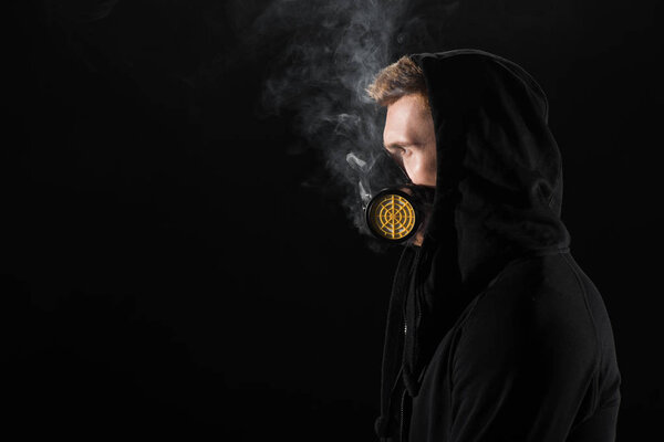 Man in black hood wearing protective filter mask surrounded by clouds of smoke