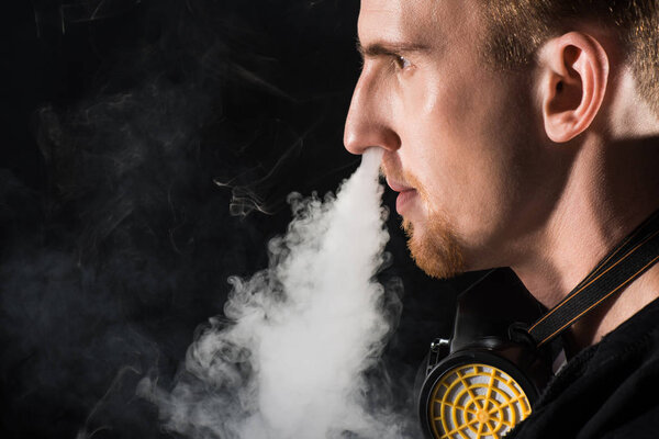 Man wearing protective filter mask exhaling smoke of electronic cigarette