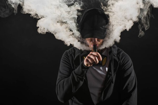 Young bearded man wearing cap exhaling smoke of electronic cigarette isolated on black