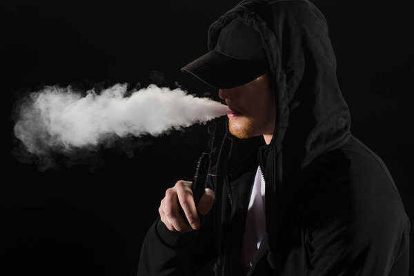 Young bearded man wearing cap exhaling smoke of electronic cigarette isolated on black