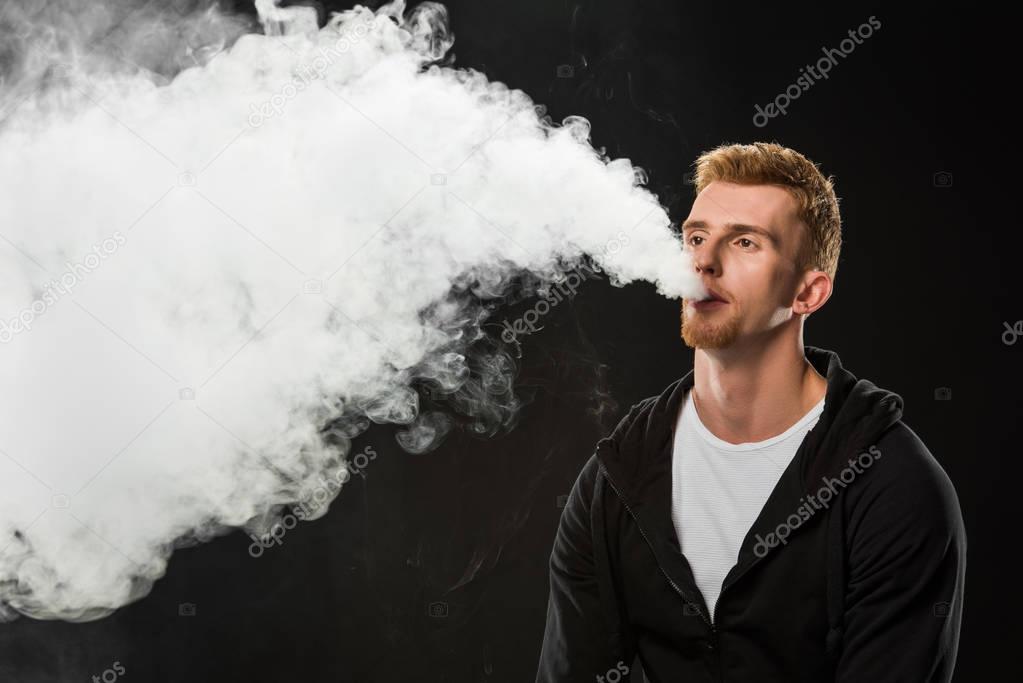 Young bearded man exhaling smoke of electronic cigarette surrounded by clouds of steam