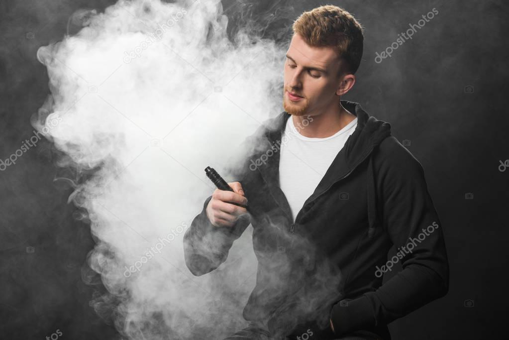 Young bearded man smoking electronic cigarette surrounded by clouds of steam