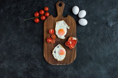 top view of arrangement of fried eggs and cherry tomatoes on wooden cutting board on dark surface clipart