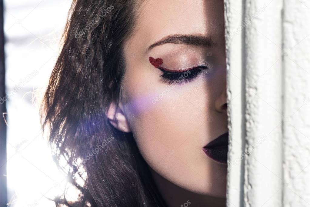 cropped image of girl with painted heart on eyelid standing with closed eyes behind column, valentines day concept  