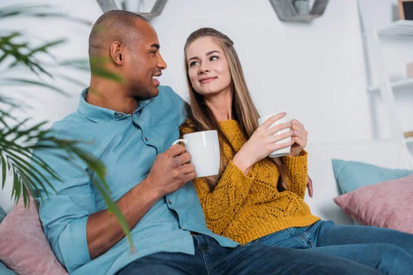 微笑的多文化夫妇坐在沙发上与咖啡 — 图库照片