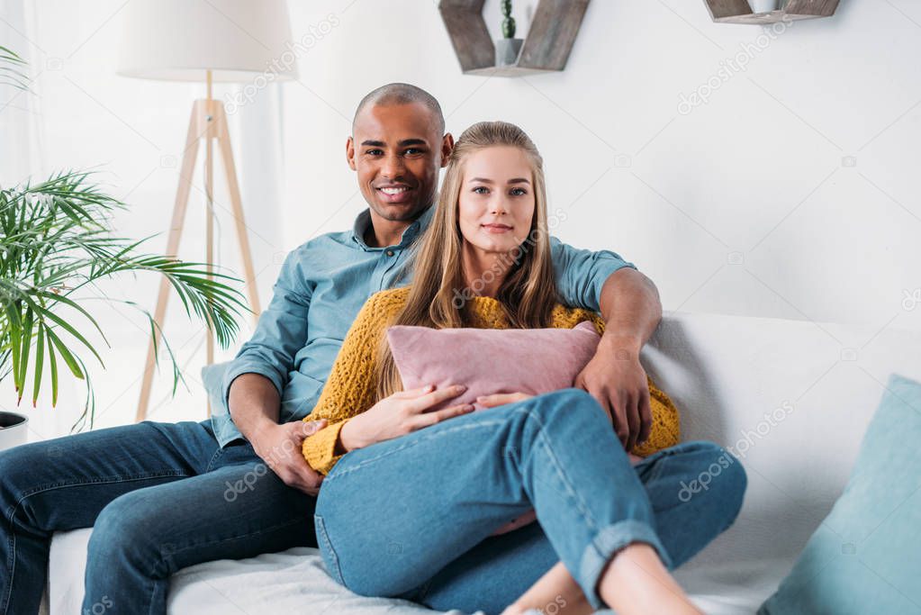 multicultural couple hugging on sofa and looking at camera