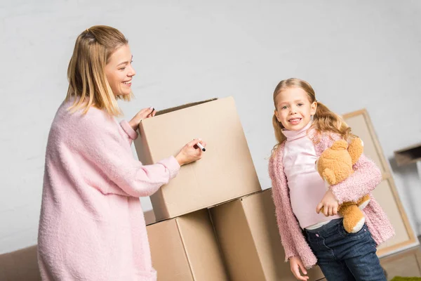 Happy Mother Daughter Teddy Bear Signing Cardboard Boxes While Relocating — Stock Photo, Image