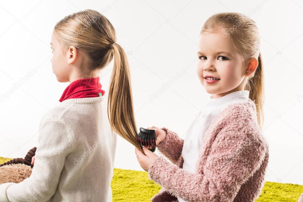 beautiful child brushing hair of sister while sitting on floor isolated on white