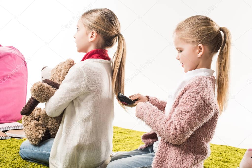 happy child brushing hair of sister while sitting on floor isolated on white