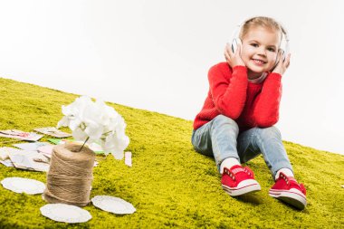 adorable little child listening music with headphones while sitting on green carpet isolated on white clipart