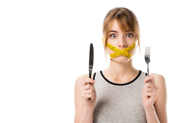 shocked young woman with measuring tape tied around her mouth holding fork and knife isolated on white
