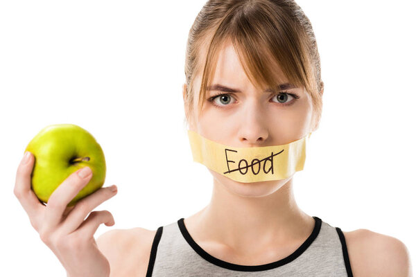 young woman with stick tape with striked through word food covering mouth holding apple isolated on white