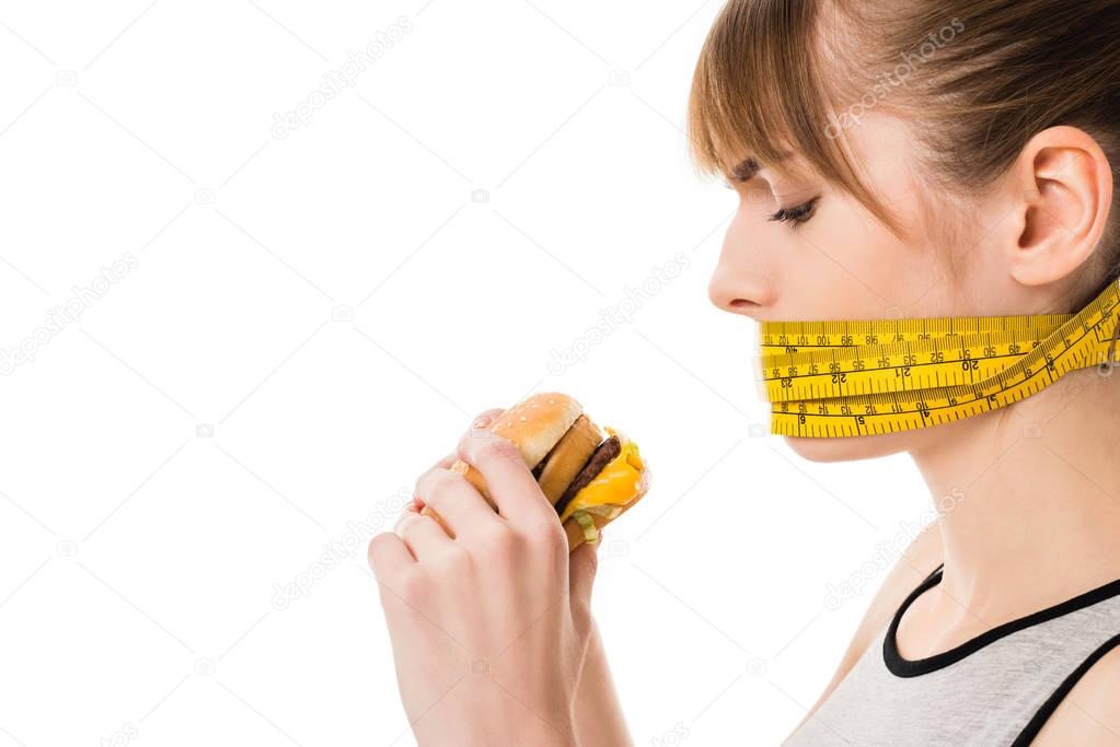 woman with mouth tied in measuring tape trying to eat burger isolated on white