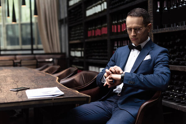 handsome man in stylish suit looking at watch while waiting for date at restaurant