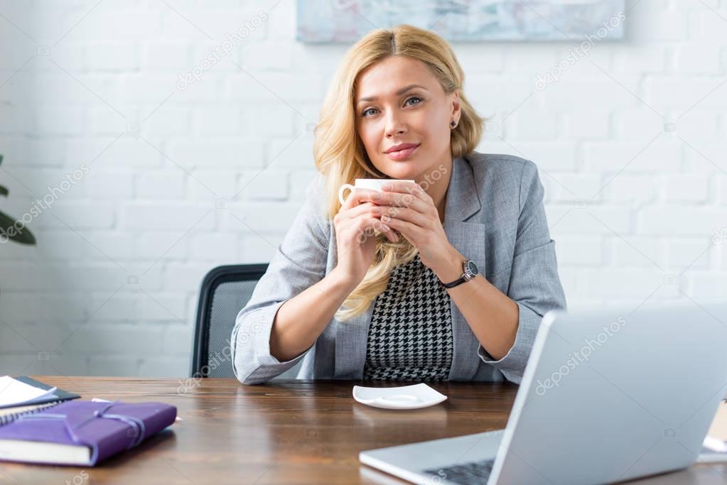 cheerful businesswoman holding cup of coffee in office and looking at camera