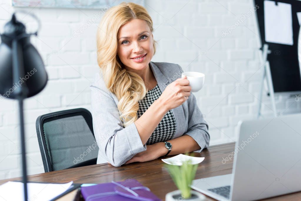 smiling businesswoman holding cup of coffee in office and looking at camera