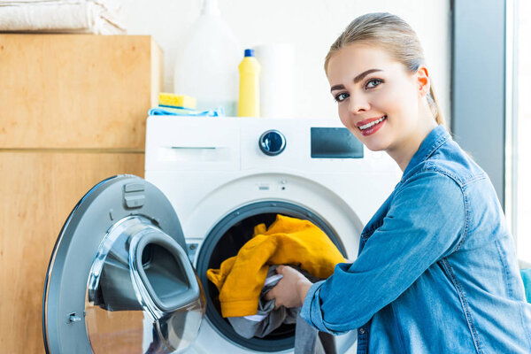 young woman smiling at camera while putting laundry into washing machine