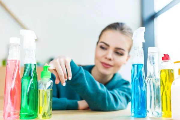 Jeune femme souriante regardant différentes bouteilles en plastique avec des produits de nettoyage — Image Jeune Femme Souriante Regardant Différentes Bouteilles Plastique Avec Des Produits Images De Stock Libres De Droits
