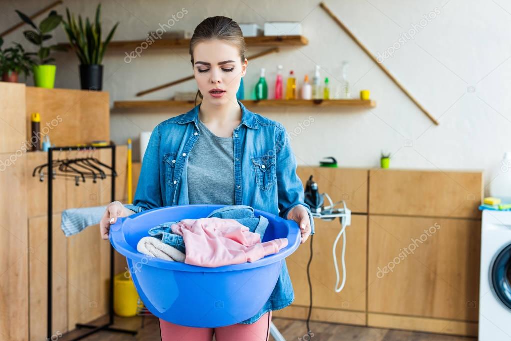 upset young woman holding plastic basin with clothes and looking at laundry