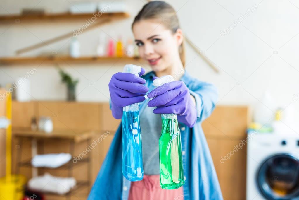 young woman in rubber gloves holding plastic bottles with cleaning fluids and smiling at camera 