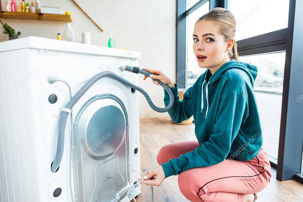 shocked young woman looking at camera while crouching near broken washing machine