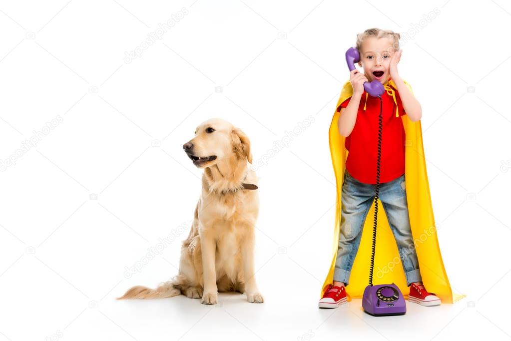 Shocked little supergirl wearing yellow cape and talking on phone with standing dog beside isolated on white