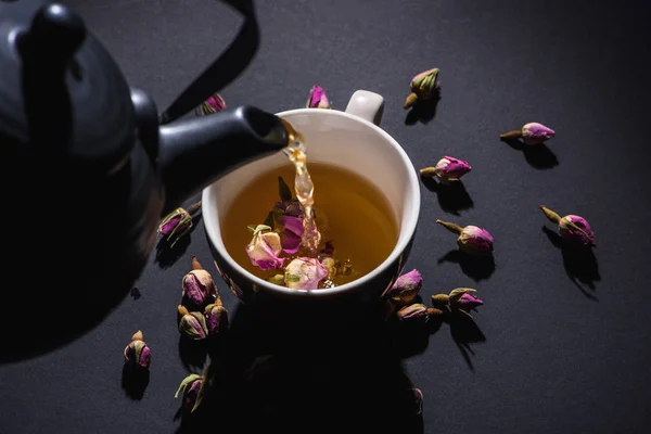 黒ポットからお茶を注ぐカップと黒地ピンク バラの蕾を乾燥 — ストック写真