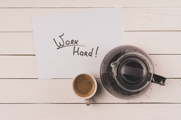 top view of cup of coffee, paper sheet with inscription work hard and coffee pot on wooden table top