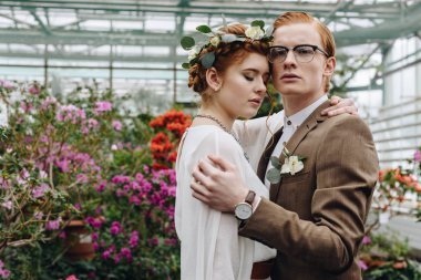 tender young red-haired wedding couple embracing while standing between flowers in botanical garden clipart