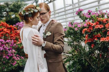 stylish young redhead wedding couple hugging between flowers in botanical garden clipart