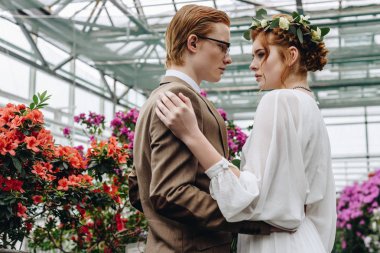 güzel genç Kızıl saçlı düğün çift kucaklayan ve birbirine Botanik bahçesinde çiçekler arasında dururken bakarak