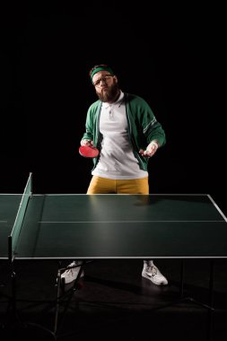 bearded sportsman with tennis equipment standing at table isolated on black clipart